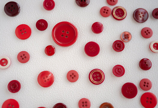 Overhead photograph showing an assortment of buttons in shades of red, maroon, and burgundy in various sizes and designs. The image highlights the contrast of sewing accessories randomly spread over a