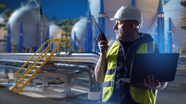 Industrial energy facility, engineer controls plant operations with a laptop and radio near storage tanks, pipelines and process equipment.