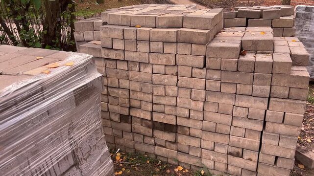 Concrete pavers stand beside a plastic wrapped stack outdoors, construction supplies, material storage, pavement renovation, autumn.