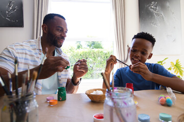 Obraz premium African American father and son painting eggs at table by window with paint pots