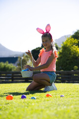 Fototapeta premium Girl child kneeling on lawn, holding white basket with colorful eggs and wearing bunny headband