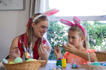 Fototapeta premium Mother and daughter sitting at home table painting eggs with brushes wearing bunny ear headbands