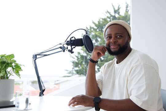 Man wearing beanie and cream T-shirt sitting at white desk recording with microphone, copy space