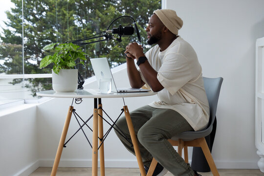 African American adult man wearing beanie recording, leaning at home studio with boom mic, laptop