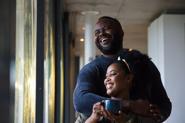 African American couple standing, embracing near tall glass windows in corridor holding blue mug © WavebreakMediaMicro