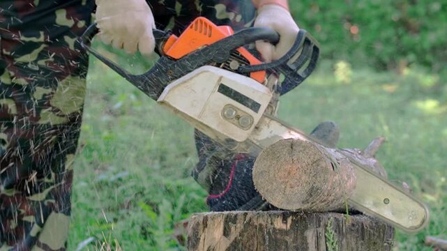 Worker in camouflage clothing cuts a log with a chainsaw, producing flying sawdust outdoors in a grassy woodland setting