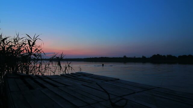 Wooden pier lake dusk. Reeds silhouette pink sunset sky, distant swimmer water.