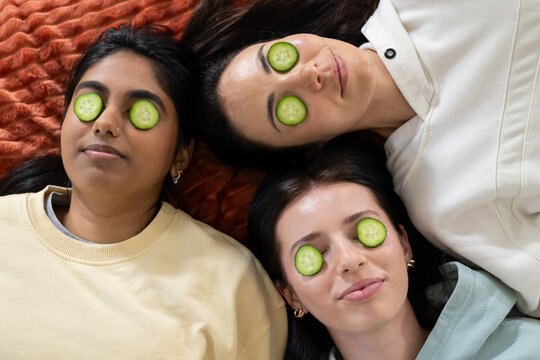 Three female friends lying on rust-orange blanket at home, wearing cucumber slices over eyes