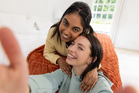 Two female friends posing and smiling while taking selfie on bed with rust blanket, smartphone