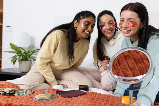 Female friends in hoodies sitting on bed applying under-eye gel patches around mirror