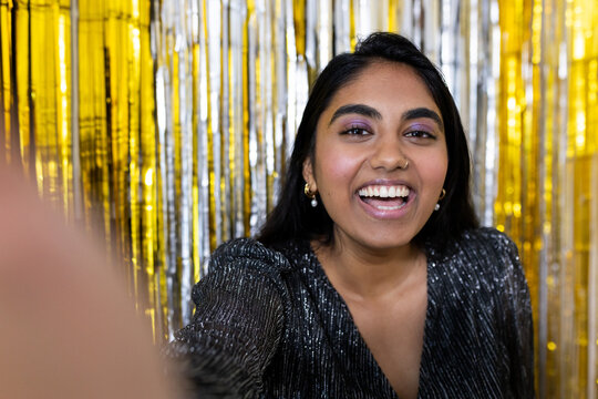 Indian woman smiling and taking selfie with smartphone in shimmery black top, metallic backdrop