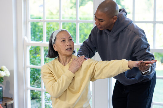 Senior Chinese woman performing arm stretch at home, African man helping near glass door with vase