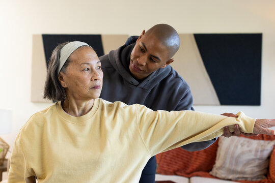 Diverse caregiver supporting senior Chinese woman wearing yellow sweatshirt extending arm at home