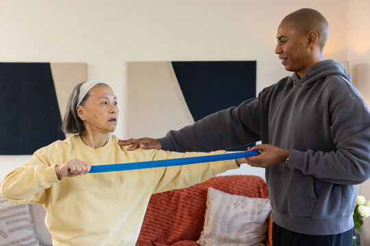 Senior Chinese woman in sweatshirt stretching blue band at home with African American man helping