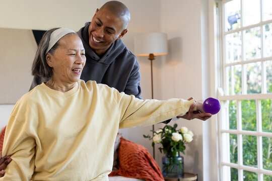 Man assisting senior Asian woman wearing yellow sweatshirt lifting purple dumbbell on sofa at home