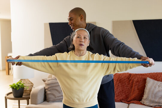 Senior Asian woman in yellow sweater stretching with blue band at home African American man guiding