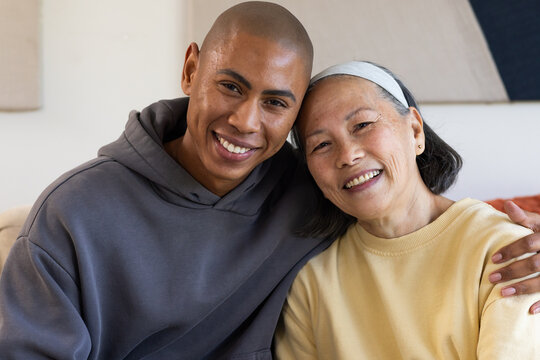 Senior Asian mother and son sitting on living room sofa, leaning in, yellow sweater, gray hoodie