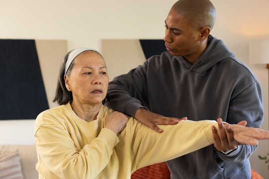 Senior Asian woman receiving guided arm stretch from African American caregiver at home on sofa