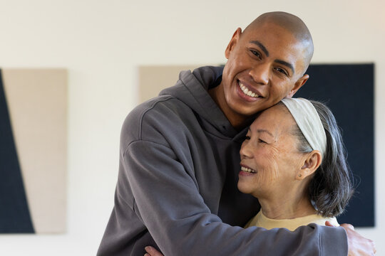 Diverse family senior mother and son embracing by pale wall with canvases wearing gray hoodie