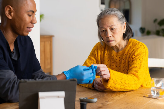 Senior Chinese woman, African-American caregiver in scrubs doing finger test home using glucometer