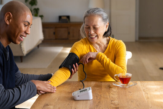 Diverse caregiver holding senior Chinese woman's arm, taking BP at table senior wearing mustard top