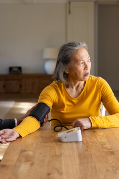 Senior Chinese woman at home wearing yellow getting blood pressure cuff adjusted by aide with meter