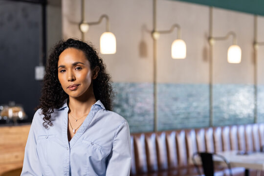 African American woman standing facing camera in cafe wearing light blue shirt near pastry dome