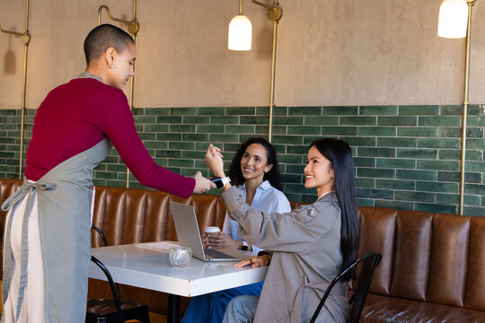 Diverse women sitting cafe table using laptop holding cup, server wearing apron handing card reader