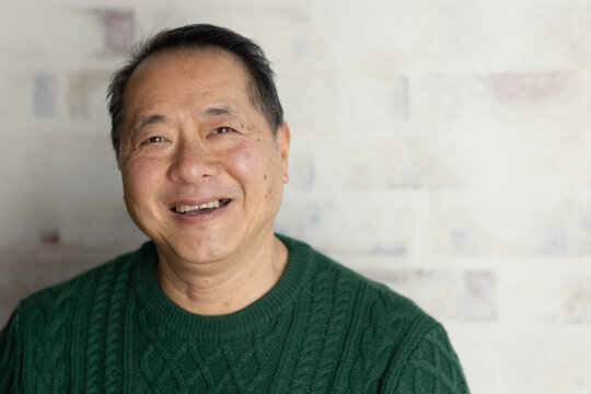 Mature Asian man smiling, sitting in studio wearing green cable-knit sweater with small framed mark