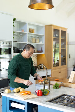 Asian mature man wearing green knit and jeans standing in kitchen feeding carrot through crank tool