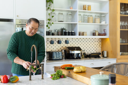 Senior Asian man in sweater rinsing greens under faucet at kitchen island, cutting board