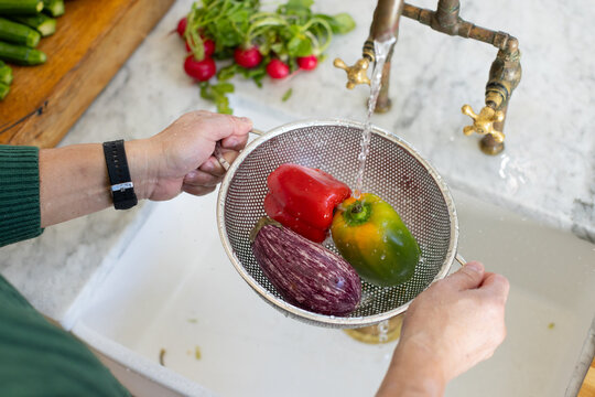 Mesh colander is rinsing peppers and eggplant under brass faucet at kitchen sink and cutting board