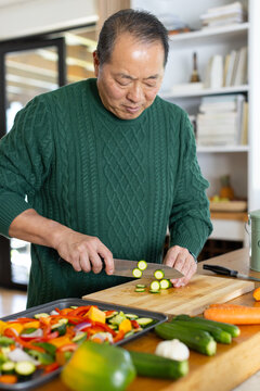 Senior Asian man in green cable-knit sweater slicing zucchini at kitchen counter with chef's knife