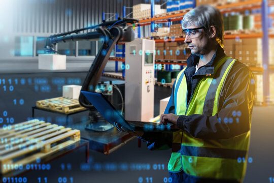Warehouse automation, engineer uses a laptop to control a robotic arm handling packages on pallets, managing automated logistics and order fulfillment operations.