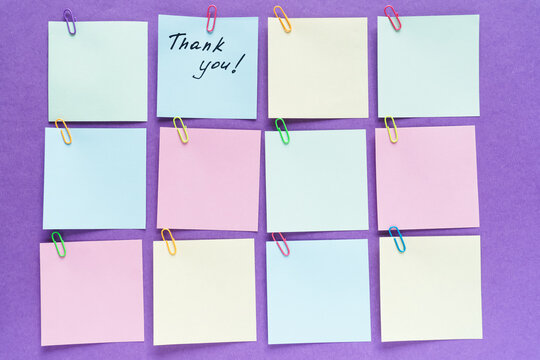 Top view flat lay horizontal photo of pastel paper notes attached with colorful paper clips in a grid pattern on purple background, with one note featuring handwritten Thank you message.