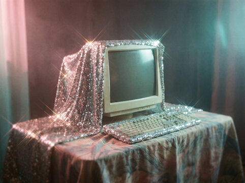 Vintage old desktop PC fully covered with sparkling silver sequins sits on dark desk under pink and cyan ambient lighting.