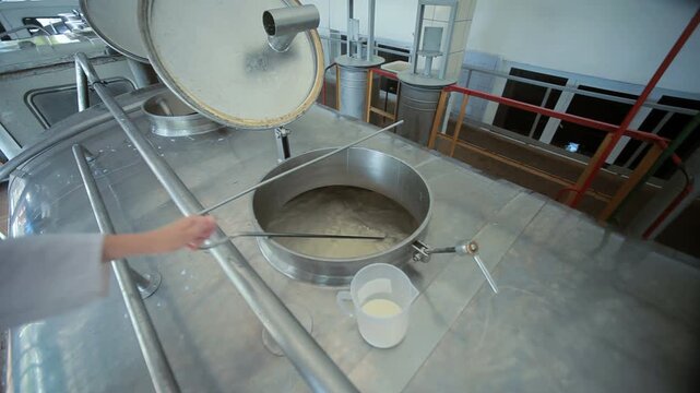 Worker stirring milk large steel vat. Dairy factory food production process.