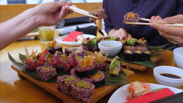 Chopsticks pick up sushi pieces from crowded serving boards during shared dining. Colorful rolls, dipping sauce, and ginger sit on table while several hands reach in with eager anticipation.