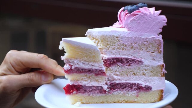 Fork lifts slice from berry layer cake on plate, revealing cream filling and soft sponge. Dessert serving continues as hand removes portion, leaving tidy edge and pastel pink topping with berries.