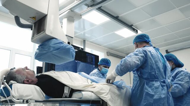 Patient lying on an angiography table while a medical team performs a procedure under C-arm X-ray guidance in a hospital cath lab.