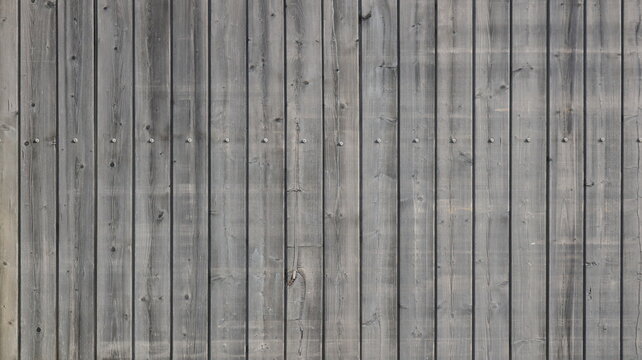 A close-up of the texture of an old vertical wooden fence. The surface consists of gray planks featuring a distinct wood grain pattern, knots, and a horizontal row of metal nails. 