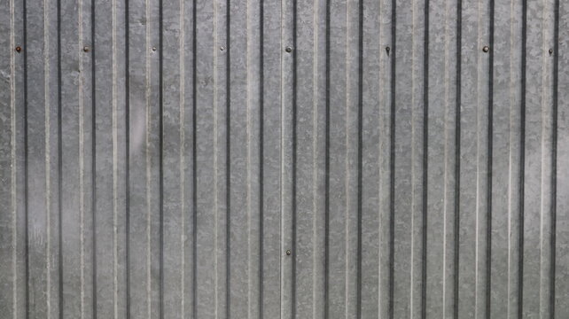 Background of an old corrugated metal sheet with a weathered texture. A grey metal wall featuring rhythmic vertical corrugations, signs of fading, minor imperfections, and visible screw heads. 