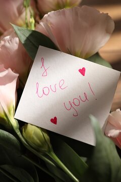 Phrase I Love You written on paper note and bouquet of eustoma flowers on table, closeup