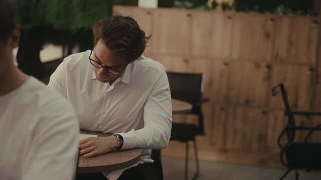 Two male students sitting at table during difficult university exam, one person struggling and asking classmate for help, who discreetly showing answers on paper. Shooting in slow motion.