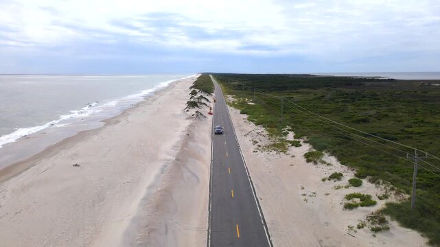 car approaches along a coastal road between dunes and Outer Banks vegetation.

