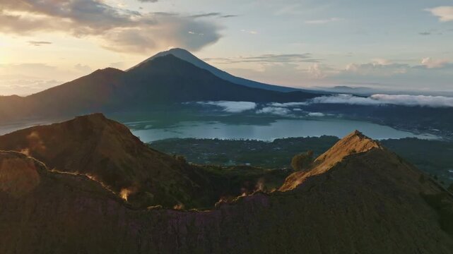Aerial sunrise over Mount Batur caldera and Lake Batur in Bali, with steaming fumarole and distant Mount Agung on the horizon, dramatic volcanic landscape under colorful sky. Drone flight panorama