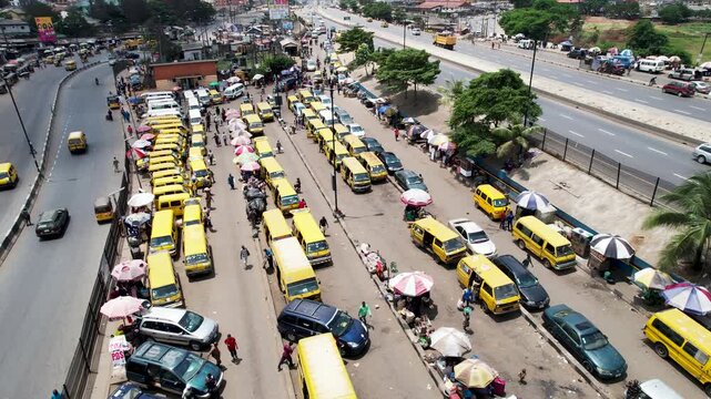 Aerial view of busy bus terminal with yellow danfo buses, commuters, and street vendors under umbrellas along the highway in Ojodu Berger, Lagos, Nigeria.