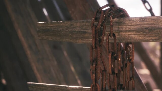 Chains are hanging loosely on a wooden post in a bright outdoor area. The chains show signs of rust and wear over time. Sunlight highlights the texture and details of the chains.