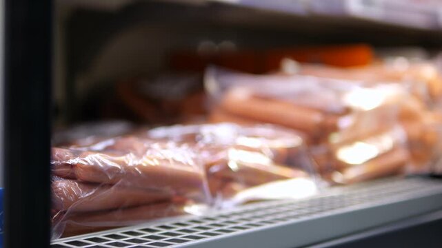 Hand selects packaged sausages from refrigerator shelf, reaching among rows of chilled meat products. Careful grocery shopping highlights convenient plastic wrapped food in store cooler.