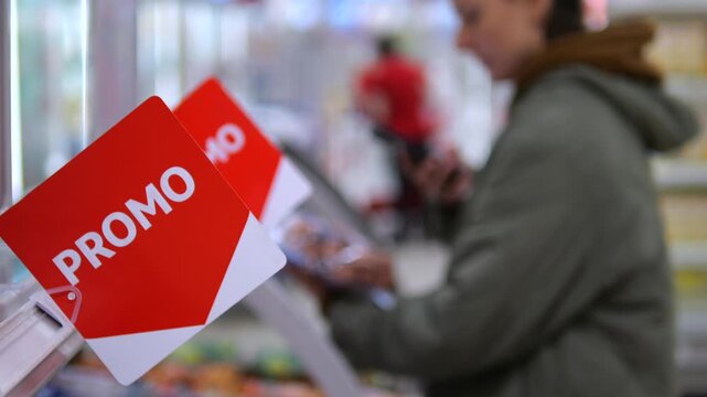 Shopper checks smartphone beside refrigerated supermarket shelves, with bright red promo tags in foreground. Customer browses chilled groceries while using mobile phone during routine store visit.